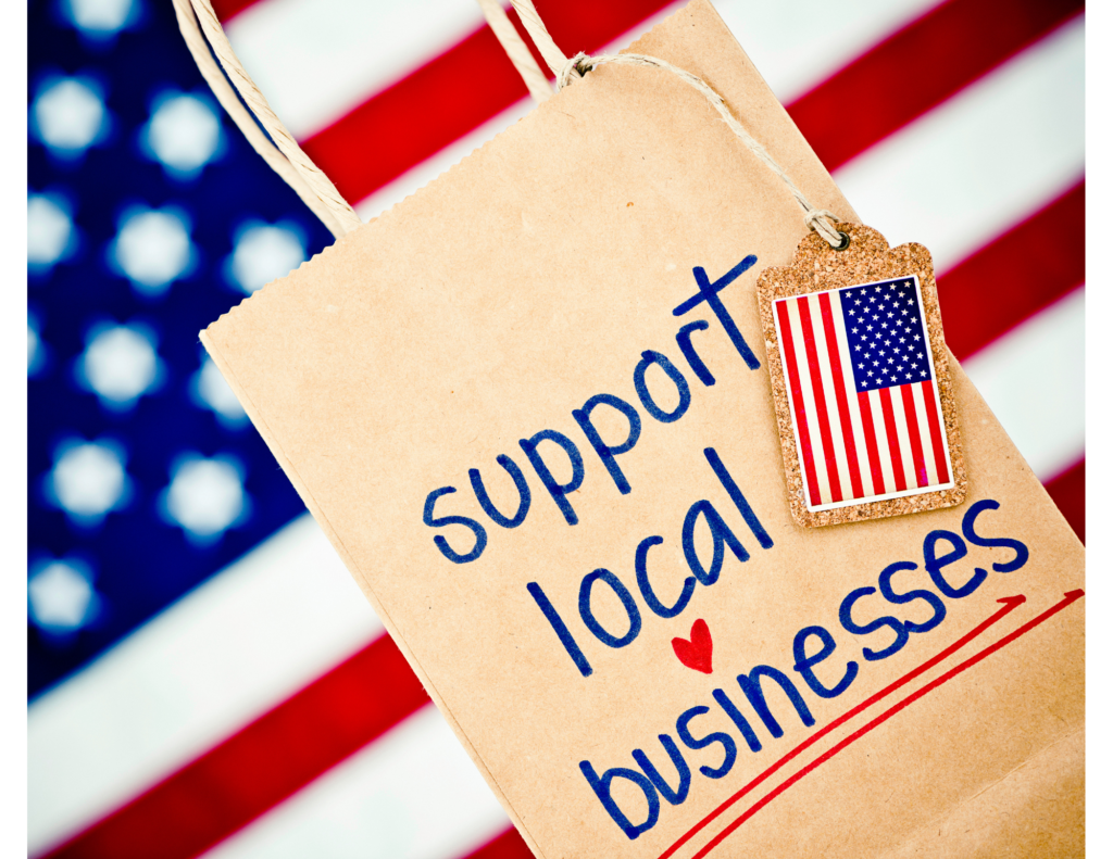 American flag as the background. There is a brown paper bag with the words support local businesses written on it in blue and red ink and a small American flag attached with brown rope to the paper bag.